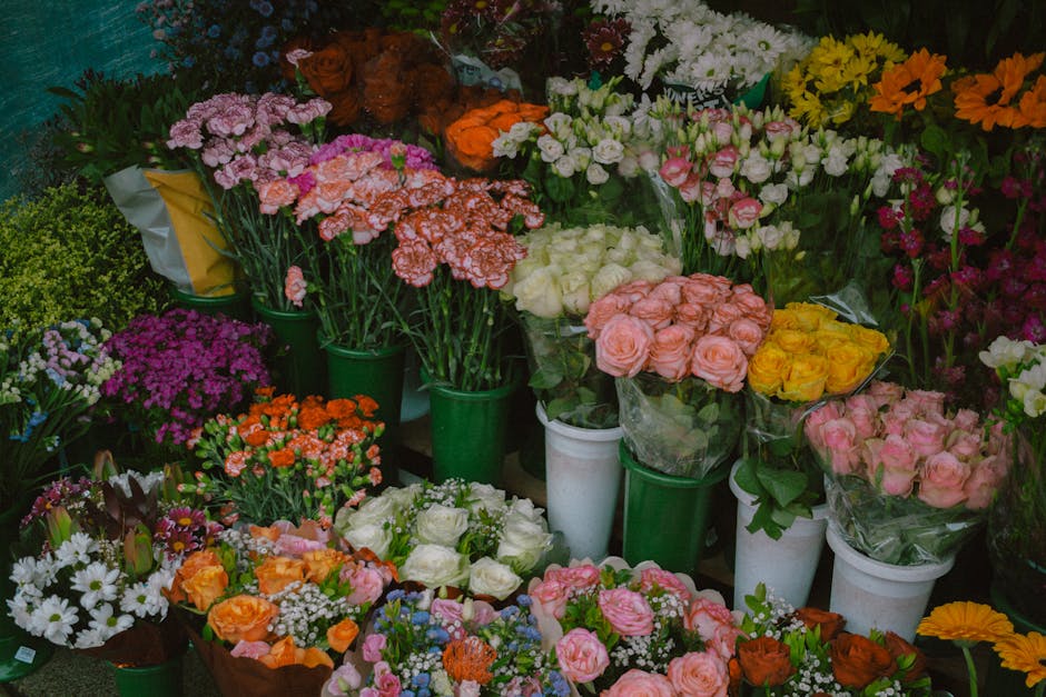 A vibrant assortment of fresh flower bouquets displayed in white and green pots at a flower shop, featuring roses in shades of pink, yellow, and peach, along with carnations, daisies, and chrysanthemu