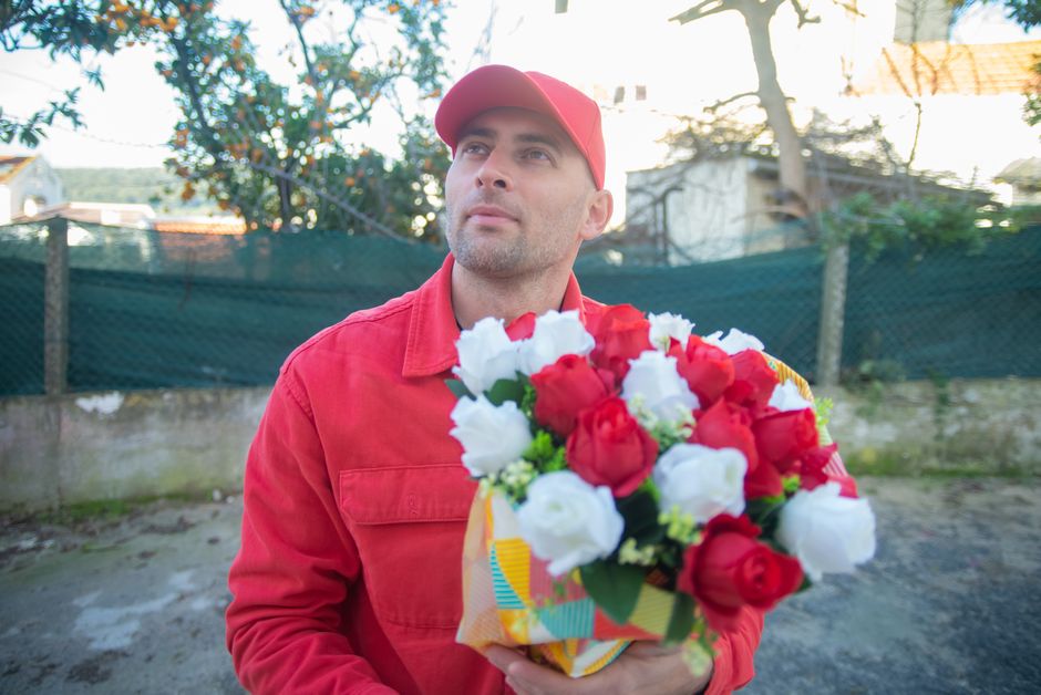 A delivery person dressed in a red uniform and matching red cap is holding a vibrant floral bouquet consisting of red and white roses, arranged in a colorful, patterned gift bag. The person stands out