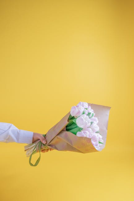 A hand holding a bouquet of fresh pink and white tulips wrapped in light brown kraft paper with a green ribbon, set against a vibrant yellow background. The tulips have smooth, slightly ruffled petals
