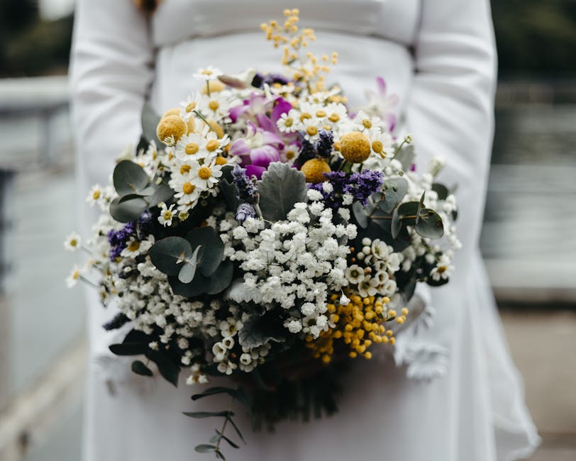 A person dressed in a white outfit holds a large, lush bouquet featuring an assortment of fresh flowers including white daisies, purple orchids, yellow mimosa, and small white baby's breath, accented 