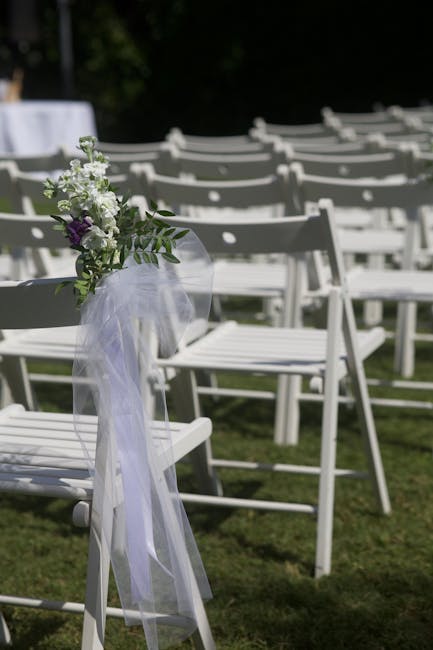 A white wooden chair at an outdoor wedding ceremony decorated with a small floral arrangement attached to the backrest, featuring white and purple flowers, green foliage, and a sheer white ribbon. The