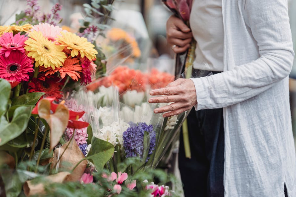 A person wearing a white long-sleeve cardigan is seen selecting a bouquet of fresh flowers at a floral market. The bouquet includes vibrant pink, yellow, and orange gerbera daisies, wrapping paper, an