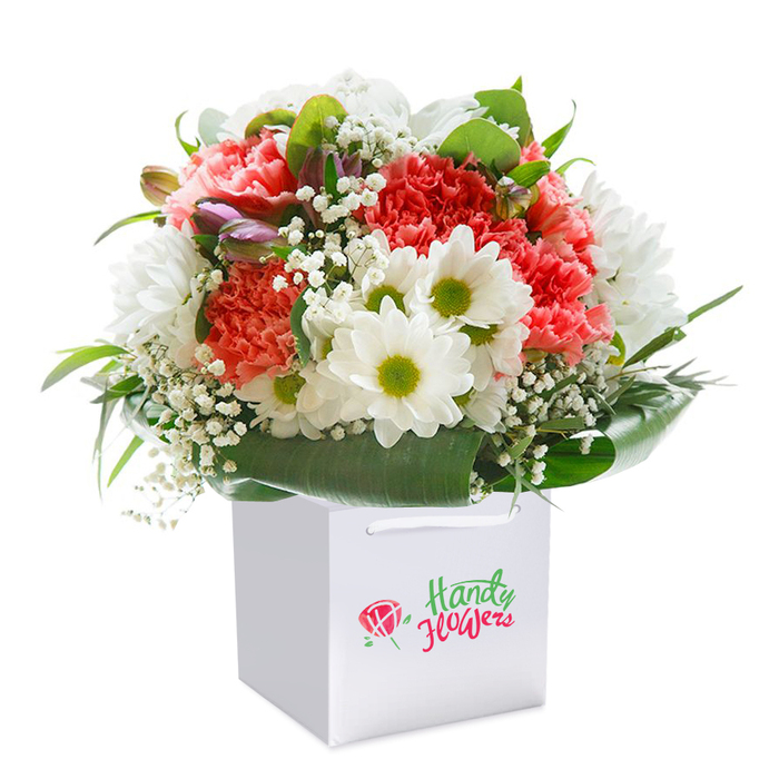 Set against a pure white background, a beautifully composed floral bouquet rises within a sleek white square gift bag, each element inspired by the charm of Crofton Park. The design features a harmonious blend of coral and blush-toned carnations, their textured petals layered in abundant ruffles. Interspersed are cheerful white daisy chrysanthemums, their luminous lime-green centers acting as small beacons of light, reminiscent of tranquil afternoons in Blythe Hill Fields. Delicate strands of baby's breath leaf through the arrangement, their softness lending grace and textural contrast. Along the left edge, closed buds tinged with purple and pink suggest the anticipation of more blooms to come, introducing depth and visual intrigue. Surrounding the bouquet, a generous sweep of glossy, deep green foliage cradles the flowers, mirroring the leafy serenity of local gardens around Crofton Park. The bag's fine ribbon handles, designed for easy portability, add an understated elegance to the presentation-ideal for thoughtful gifting or doorstep delivery. Meticulously rounded in silhouette, the bouquet embodies freshness, abundance, and a sense of gentle celebration, making it a heartfelt choice for marking birthdays, anniversaries, or simply showing care in the Crofton Park community.