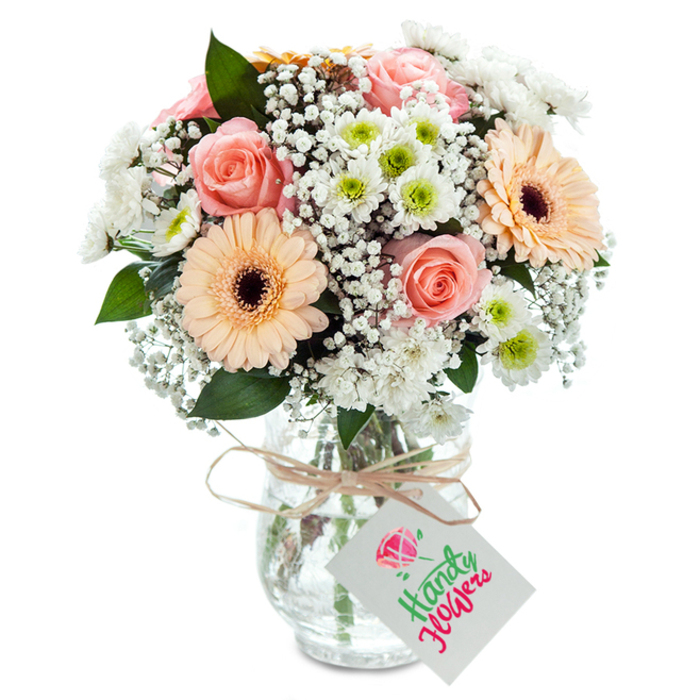 This radiant bouquet, freshly arranged for Crofton Park, stands proudly in a clear glass vase against a pristine white background. Soft, plush roses with peach-pink tones form a romantic heart, while two peach-toned gerbera daisies with expressive brown centers bring confident vibrancy. Tucked among the signature blooms, clusters of delicate baby's breath and white chrysanthemums-each one crowned with a bright green center-harmonize for a light, airy feel, reminiscent of the open parks and tree-lined streets surrounding Crofton Park Station. Sparse stems of eucalyptus weave throughout, breaking up the pastel palette with mossy green depth and subtle fragrance. The whole arrangement is gathered in a clear, slightly ribbed glass vase whose visible stems and water signal both freshness and craftsmanship. Around the neck, a simple raffia ribbon ties off a white rectangular tag, ready for a handwritten message-a small gesture that can brighten a friend's day from Honor Oak to Brockley. This bouquet, awash in gentle natural light, offers serenity and style to any occasion throughout the SE4 community.